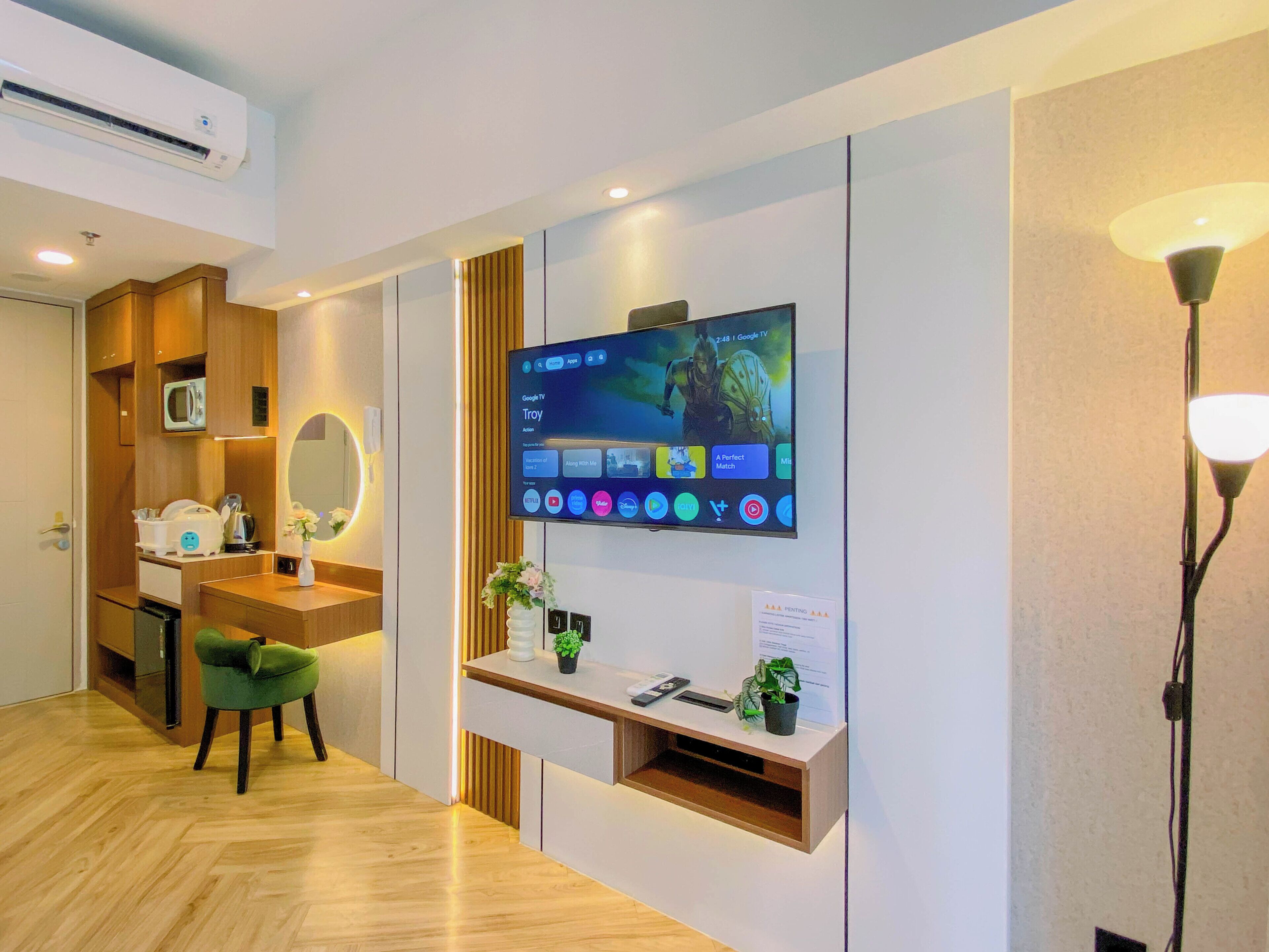 A hotel room with a flat-screen TV mounted on the wall, a wooden vanity with a mirror, a green chair, and a small shelf with a plant.