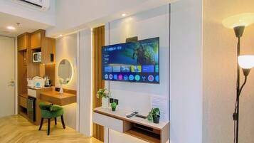 A hotel room with a flat-screen TV mounted on the wall, a wooden vanity with a mirror, a green chair, and a small shelf with a plant.