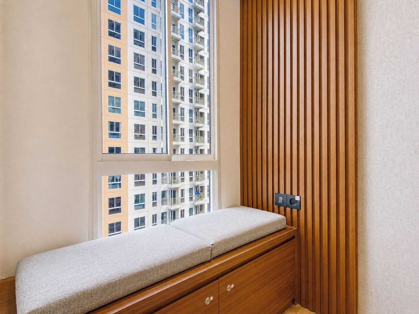 A room with a built-in bench, a window overlooking a high-rise building, and a wooden slat wall.