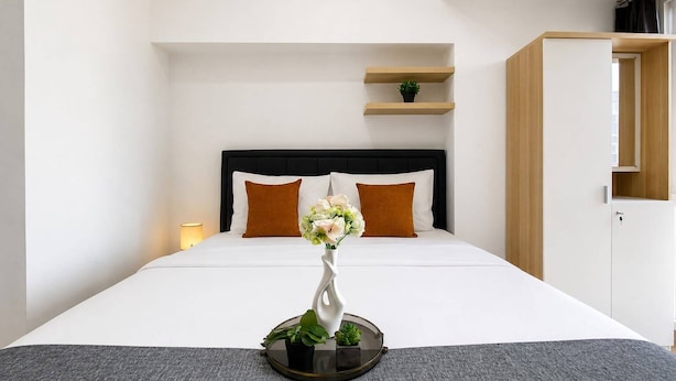 A hotel room with a bed, a bedside lamp, a small shelf with a plant, and a wardrobe. - Modern Well Located Studio Room At Apartment Tokyo Riverside Pik 2 (Jakarta)