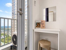 Un balcón con vistas a un paisaje urbano, una consola blanca con un espejo y dos libros, y un taburete color beige.