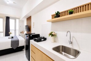 A modern hotel room with a bed, a flat-screen TV, a coffee machine, a sink, and a wooden shelf with plants. - Modern Well Located Studio Room At Apartment Tokyo Riverside Pik 2 (Jakarta)
