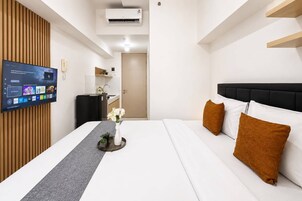 Habitación de hotel con cama, televisor montado en la pared, una kitchenette pequeña y una puerta que da a otra habitación.