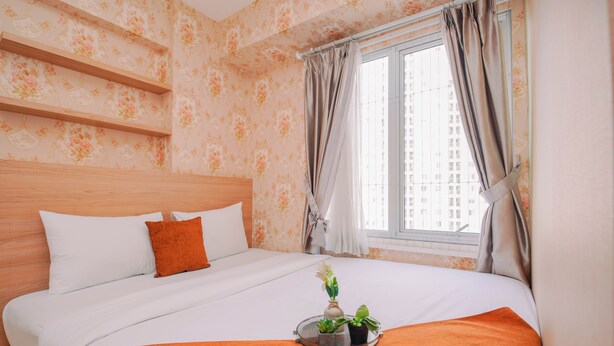 Una cama bien hecha con ropa de cama blanca y naranja, una mesita de noche con una bandeja de plantas y una ventana con cortinas.