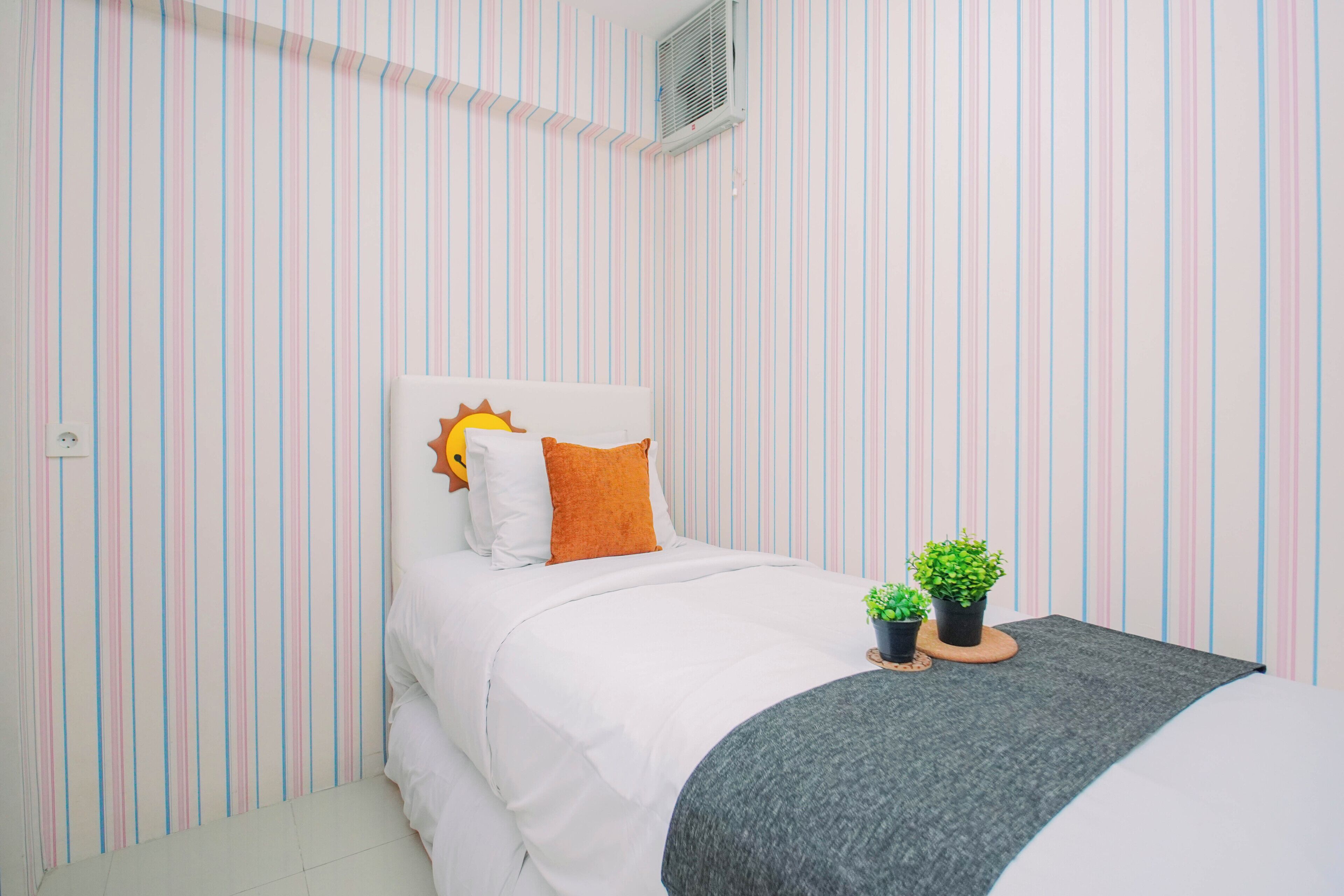 Un dormitorio con un estampado de rayas en la pared, una cama con ropa de cama blanca, un cojín con forma de sol, dos plantas pequeñas en macetas sobre una mesita de noche y una manta gris.
