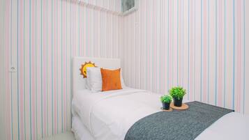 A bedroom with a striped wall pattern, a bed with white bedding, a sun-shaped pillow, two small potted plants on a bedside table, and a grey throw blanket.