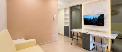 A modern living room with a sofa, a dining table with chairs, a flat-screen TV mounted on the wall, and a built-in shelving unit.