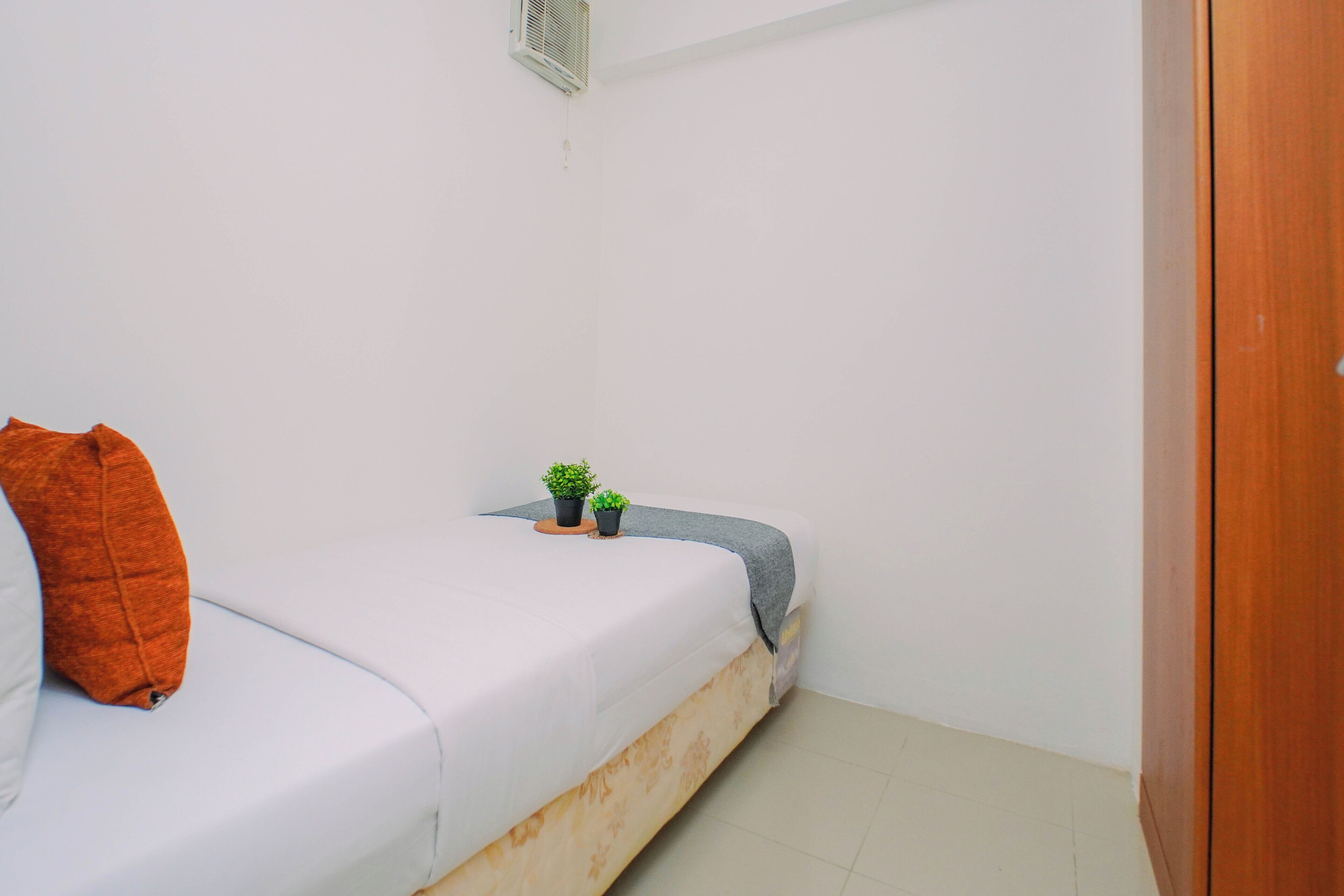 Una piccola stanza con un letto singolo, una porta in legno, un materasso bianco, un cuscino marrone e due piccoli vasi con piante su un sottobicchiere di legno.