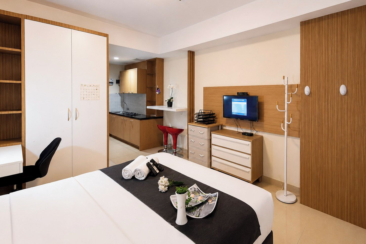 Quarto de hotel com cama, escrivaninha, televisão, guarda-roupa e cozinha compacta.