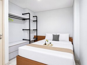 Quarto de hotel moderno com cama, cabeceira de madeira, mesa de cabeceira e prateleira embutida com cabides.