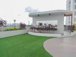 A modern outdoor area with artificial grass, planters with various plants, and a white structure with a glass window. - Cozy Compact 2Br At Apartment Patraland Urbano (Bekasi)