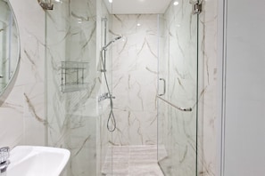 Un baño moderno con ducha envuelta en vidrio, paredes de mármol y un lavabo blanco.