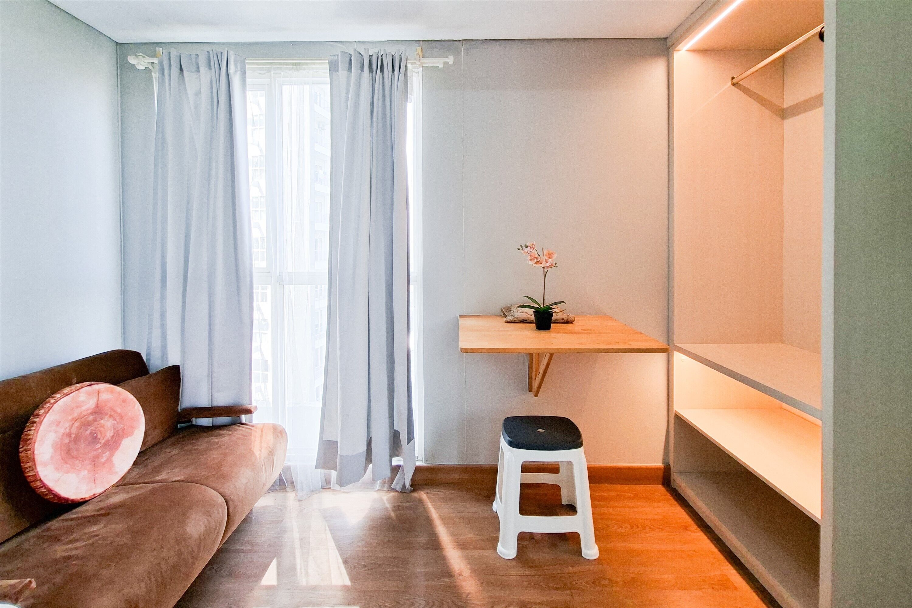 Un espacio habitable pequeño con un sofá marrón, un taburete blanco, un estante de madera y una ventana con cortinas traslúcidas.
