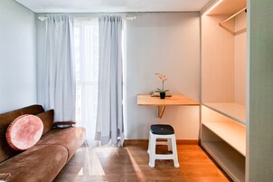Un espacio habitable pequeño con un sofá marrón, un taburete blanco, un estante de madera y una ventana con cortinas traslúcidas.
