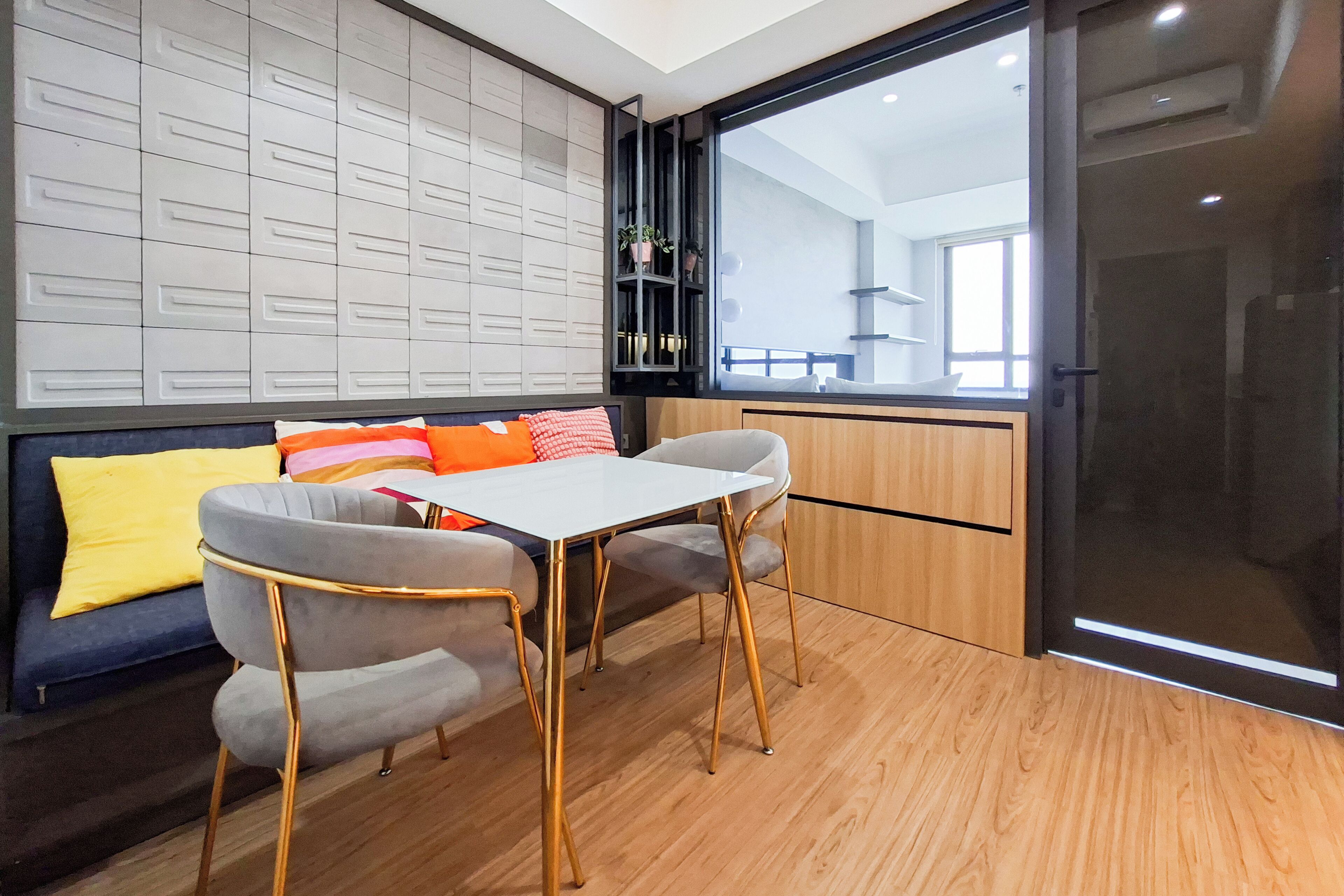 Un interior moderno con piso de madera, una mesa blanca, dos sillas grises, un sofá con cojines coloridos y un ventanal con vistas al exterior.
