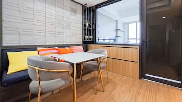 Un interior moderno con piso de madera, una mesa blanca, dos sillas grises, un sofá con cojines coloridos y un ventanal con vistas al exterior.
