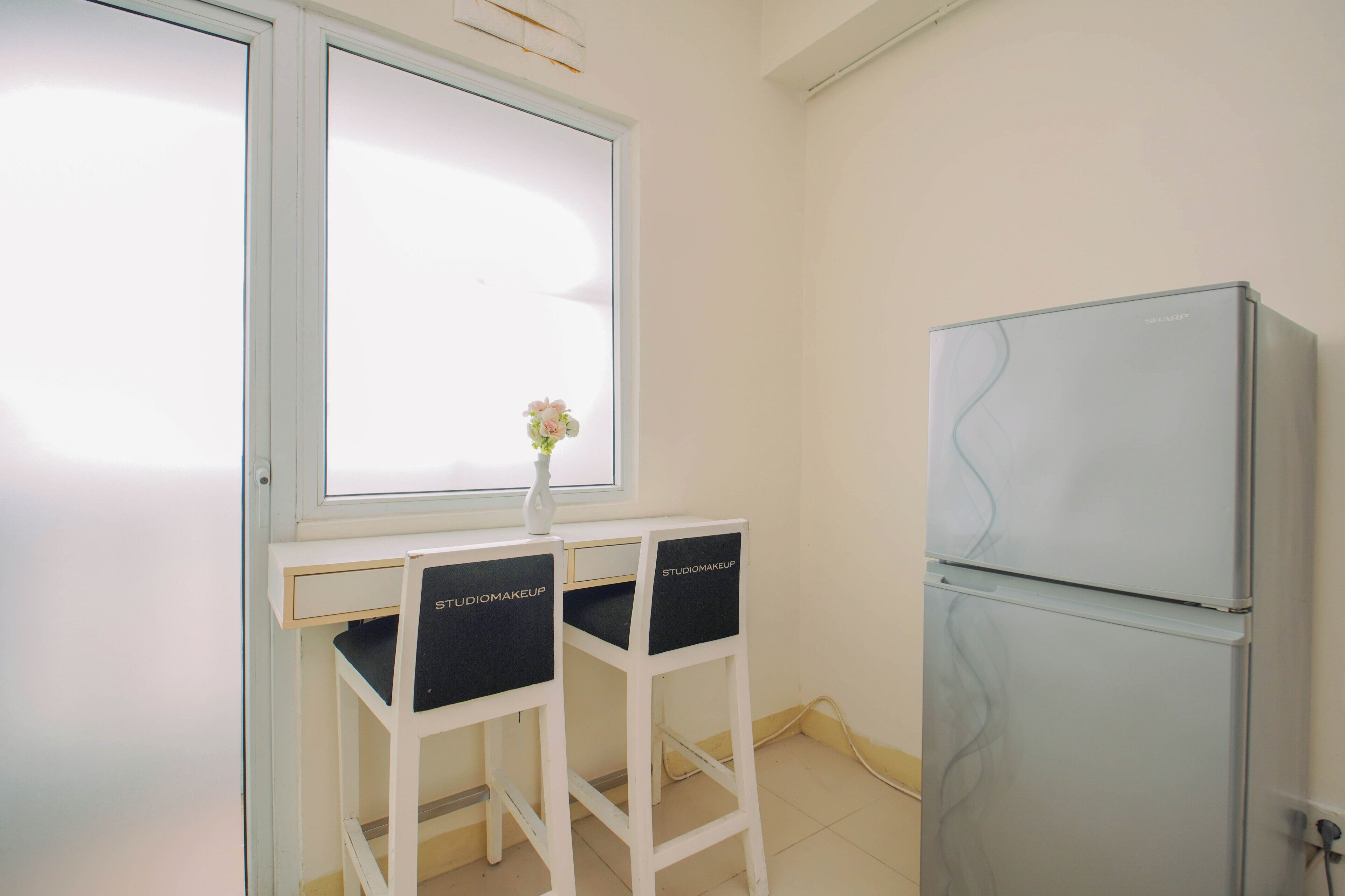 Een kamer met een wit bureau, twee zwarte opbergboxen met de tekst 'STUDIOMAKEUP', een witte koelkast en een raam met een witte luik.