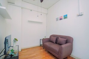 A modern living room with a wooden floor, a purple sofa, a small side table, and a flat-screen TV. - Cozy Minimalist 1Br At Apartment Green Pramuka City (Jakarta)