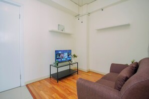 A living room with a flat-screen TV on a glass table, a brown sofa, wooden flooring, and white walls. - Cozy Minimalist 1Br At Apartment Green Pramuka City (Jakarta)