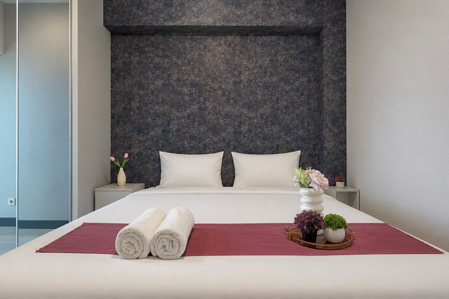 Habitación de hotel con una cama grande, dos almohadas, un plaid rojo, un florero con flores y una pequeña planta en una mesita de noche.