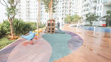 Un parque infantil con tobogán y estructuras para escalar, rodeado de palmeras y un edificio residencial al fondo.