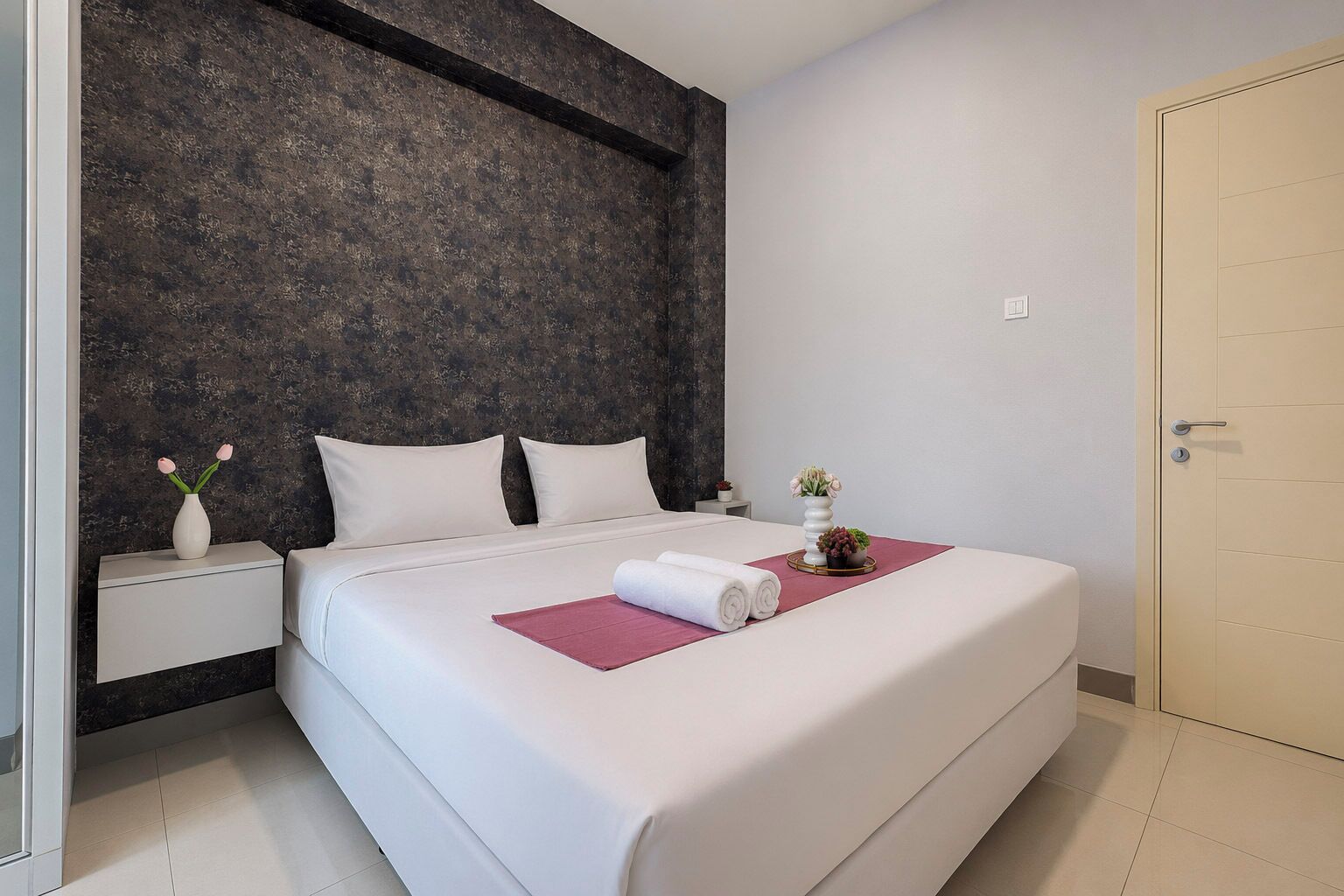 A modern hotel room with a large bed, a dark textured wall, a white door, and a small bedside table with a vase of flowers.