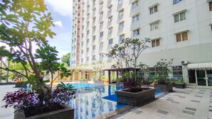 A hotel courtyard with a swimming pool, potted plants, and a tall building in the background. - Sleek Modern Studio At Apartment Pinewood (Sumedang)