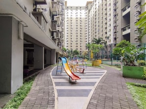 A playground with slides and swings, surrounded by a paved walkway and greenery, in a residential area with high-rise buildings. - Warm Homey 2Br At Apartment Gateway Ahmad Yani Cicadas (Bandung)