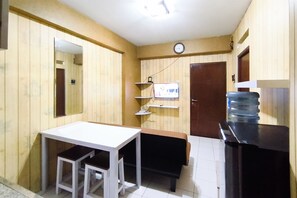Une petite pièce avec une table blanche, un banc, une horloge au mur et un distributeur d’eau.