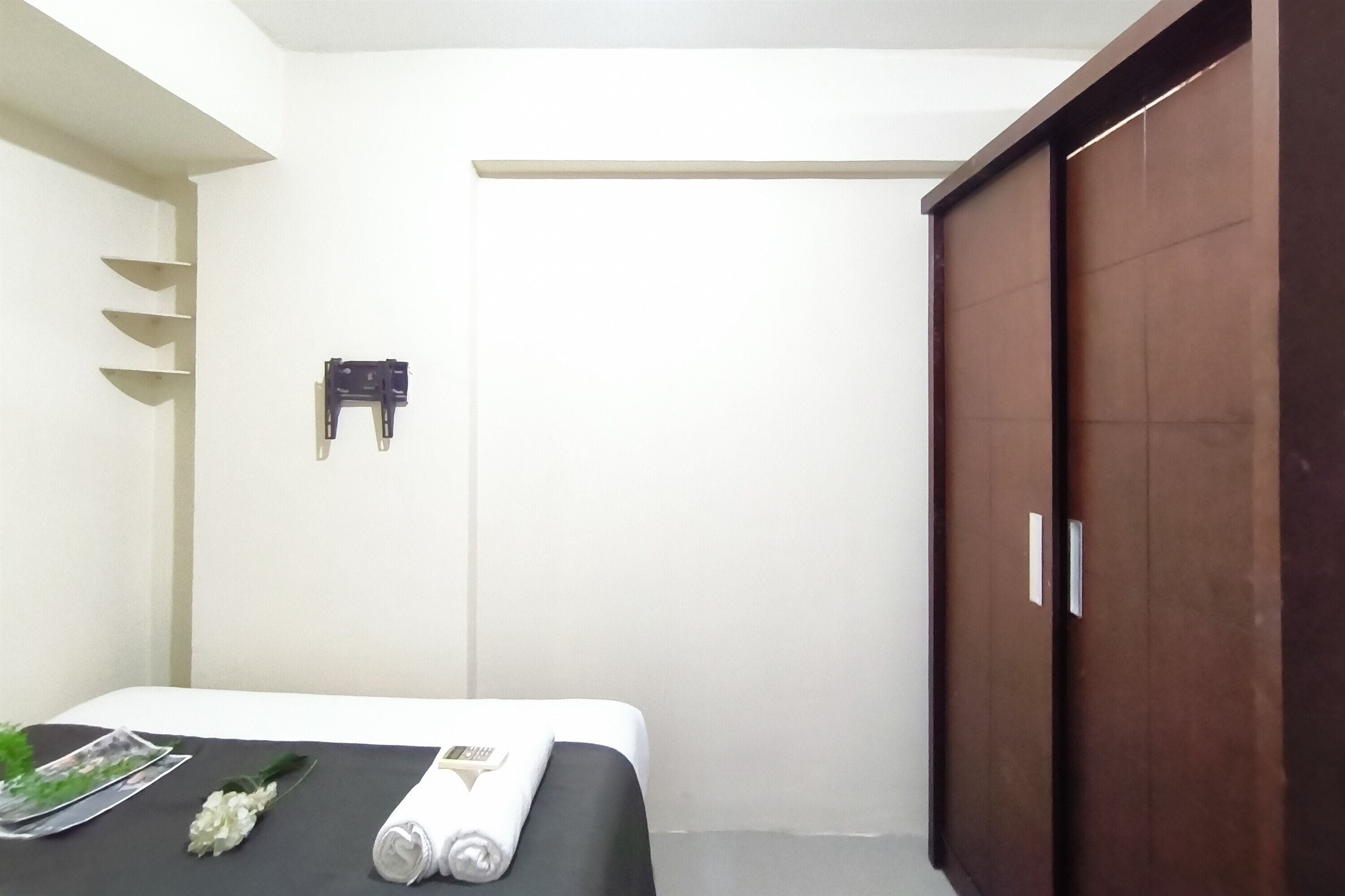 Une chambre d’hôtel avec un lit, une armoire en bois, un interrupteur fixé au mur et une petite étagère sur laquelle se trouve un objet décoratif.