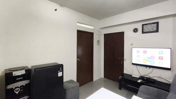A room with a flat-screen TV mounted on the wall, a black entertainment unit, a grey ottoman, and a door leading to another room. - Comfortable 2Br At Apartment Gateway Ahmad Yani Cicadas (Bandung)