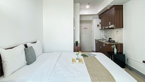 A hotel room with a bed, a bedside table with a tray, a small potted plant, and a view into the adjacent kitchen area. - Comfortable And Minimalist Studio Room At Apartment Grand Kamala Lagoon (Bekasi)