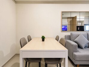 A modern living room with a grey sofa, a wooden dining table with six chairs, and a small potted plant. - Modern Warm 2Br At Apartment Meikarta District 2 (Cikarang)