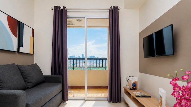 En moderne stue med en grå sofa, et fladskærms-tv, et træsidebord med en vase med lyserøde blomster, og en skydedør til en balkon med udsigt over byen.