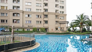 En swimmingpool med blå ternede fliser, omgivet af en lav murstensmur. I baggrunden et fleretasagers byggeri med altaner og vinduer.