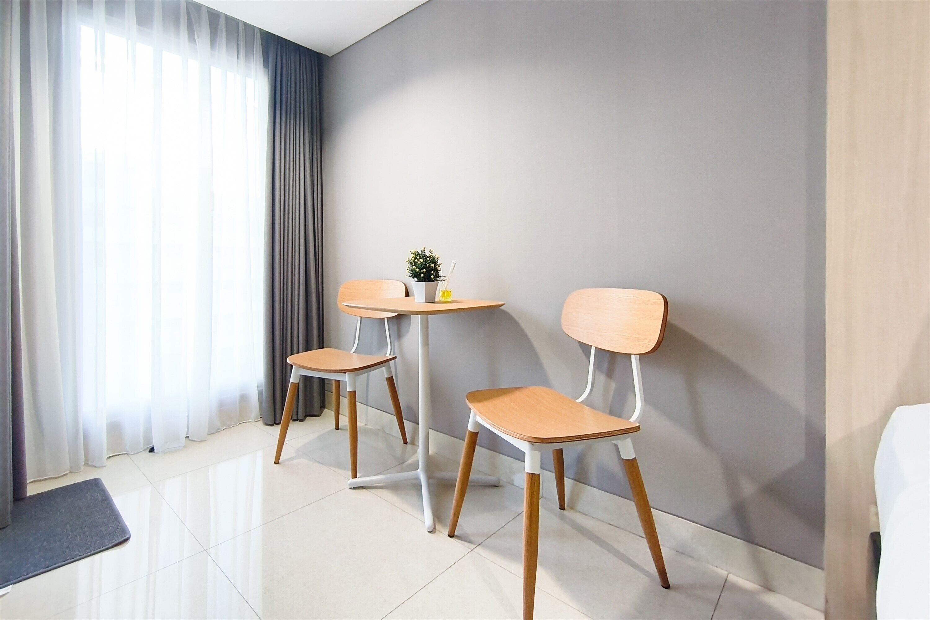 A modern hotel room with a small round table and two chairs, a potted plant, and a view of the outside through sheer curtains.