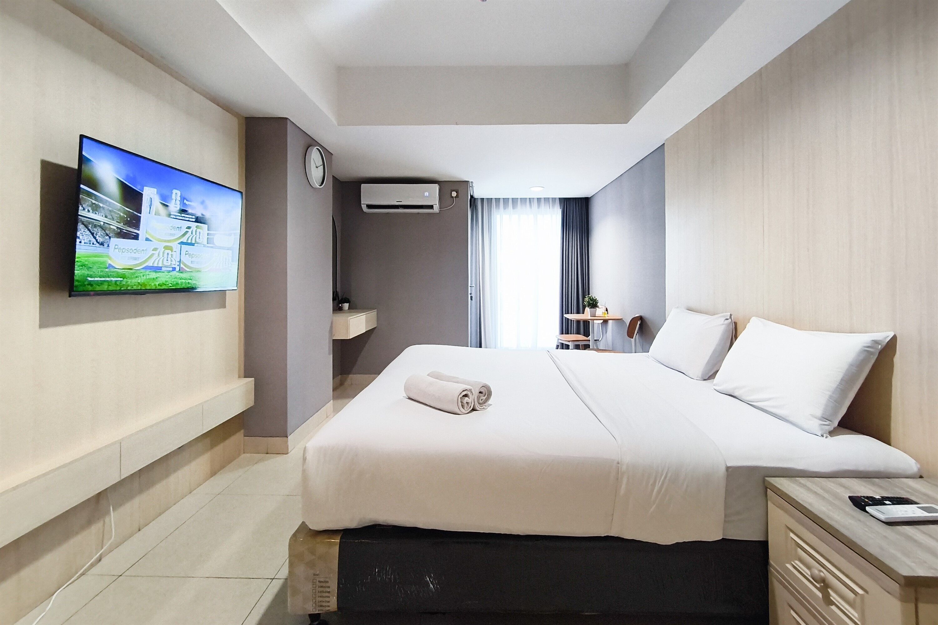 A hotel room with a bed, a TV mounted on the wall, a clock, an air conditioning unit, a small table with a chair, and a bedside table with a phone and a calculator.