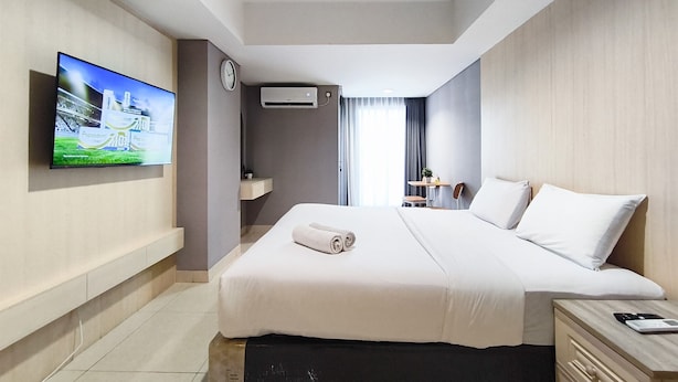 Quarto de hotel com cama, televisão na parede, relógio, aparelho de ar condicionado, pequena mesa com cadeira e mesa de cabeceira com telefone e calculadora.