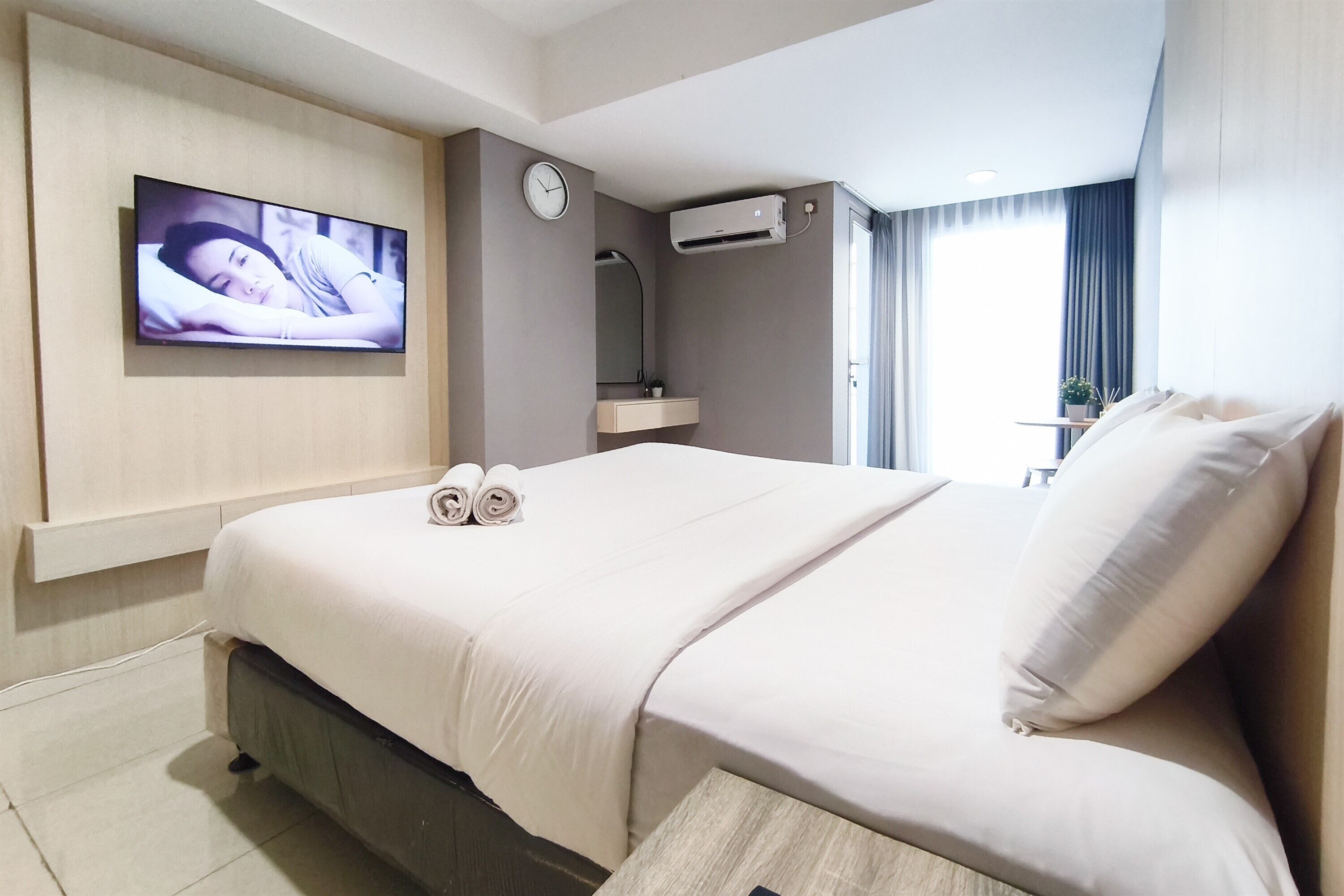 A hotel room with a bed, a TV mounted on the wall, a clock, and an air conditioning unit.