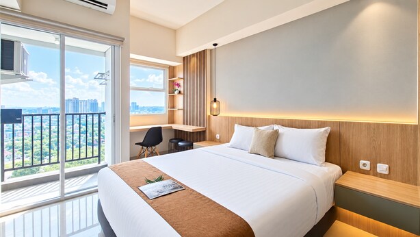 A modern hotel room with a large bed, a desk, and a balcony with a city view. - Neat And Tidy Studio At Apartment Parkland Avenue (Tangerang)