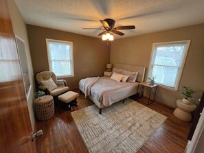 A bedroom with a bed, a chair, a bedside table, a ceiling fan, and two windows with blinds.