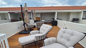 Una terraza en la azotea con sillones de descanso y sombrillas, suelo de madera y tejado de tejas rojas.