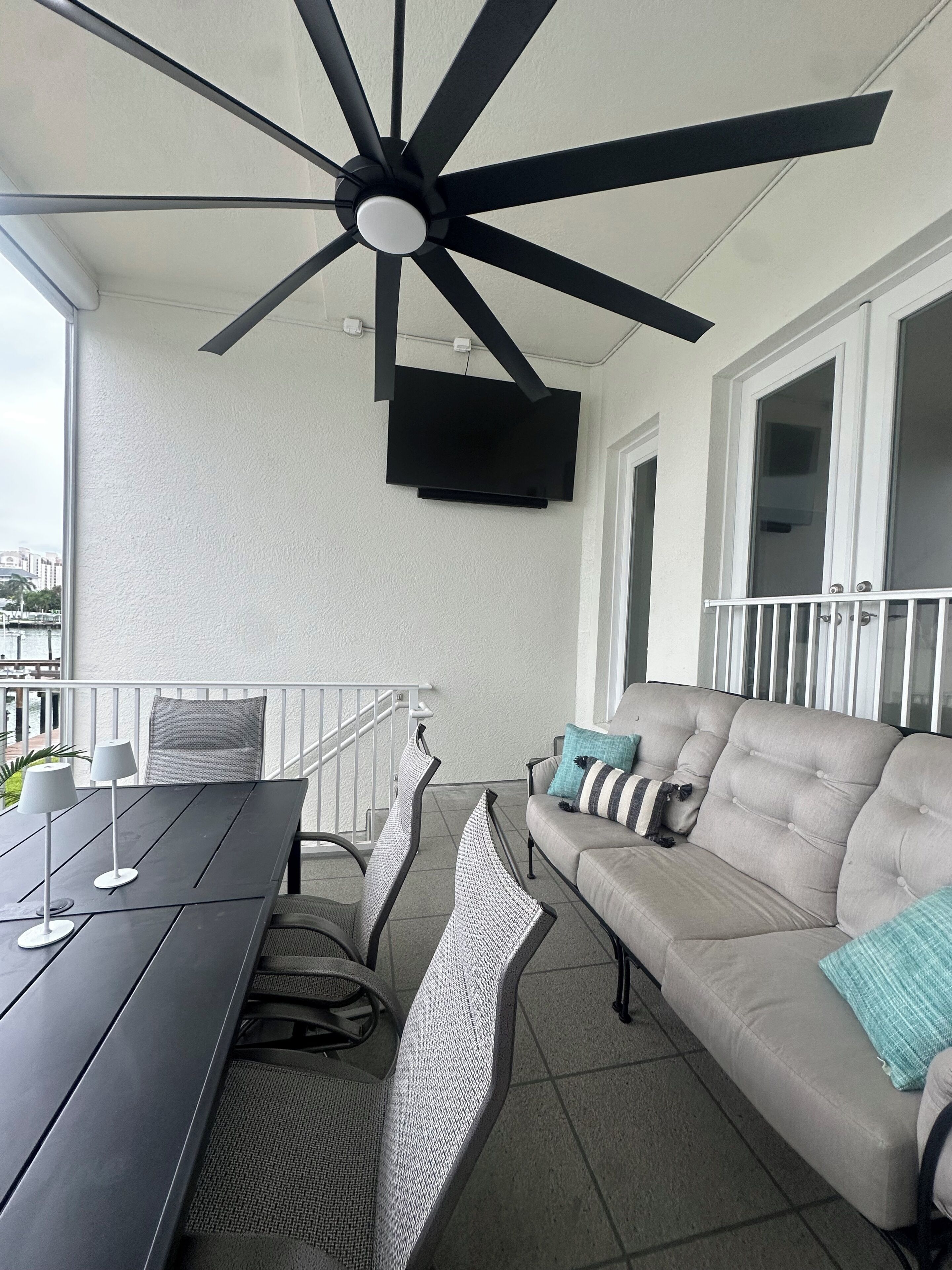 Un balcon avec un ventilateur de plafond, une table avec des chaises, un canapé avec des coussins et un téléviseur fixé au mur.