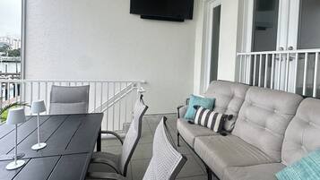 A balcony with a ceiling fan, a table with chairs, a sofa with cushions, and a mounted TV.
