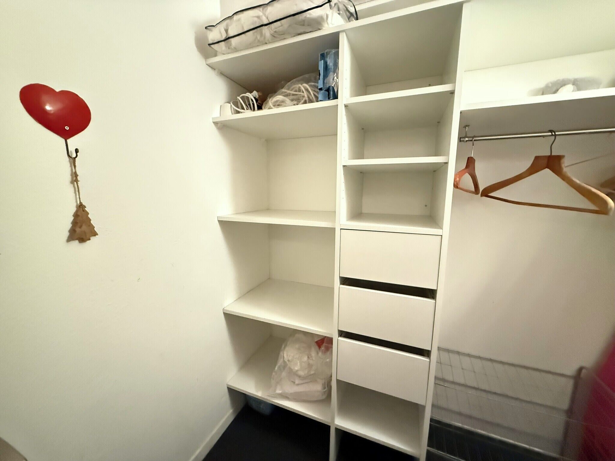 A white closet with multiple shelves and hanging space, a red heart-shaped balloon, and a small Christmas tree decoration.