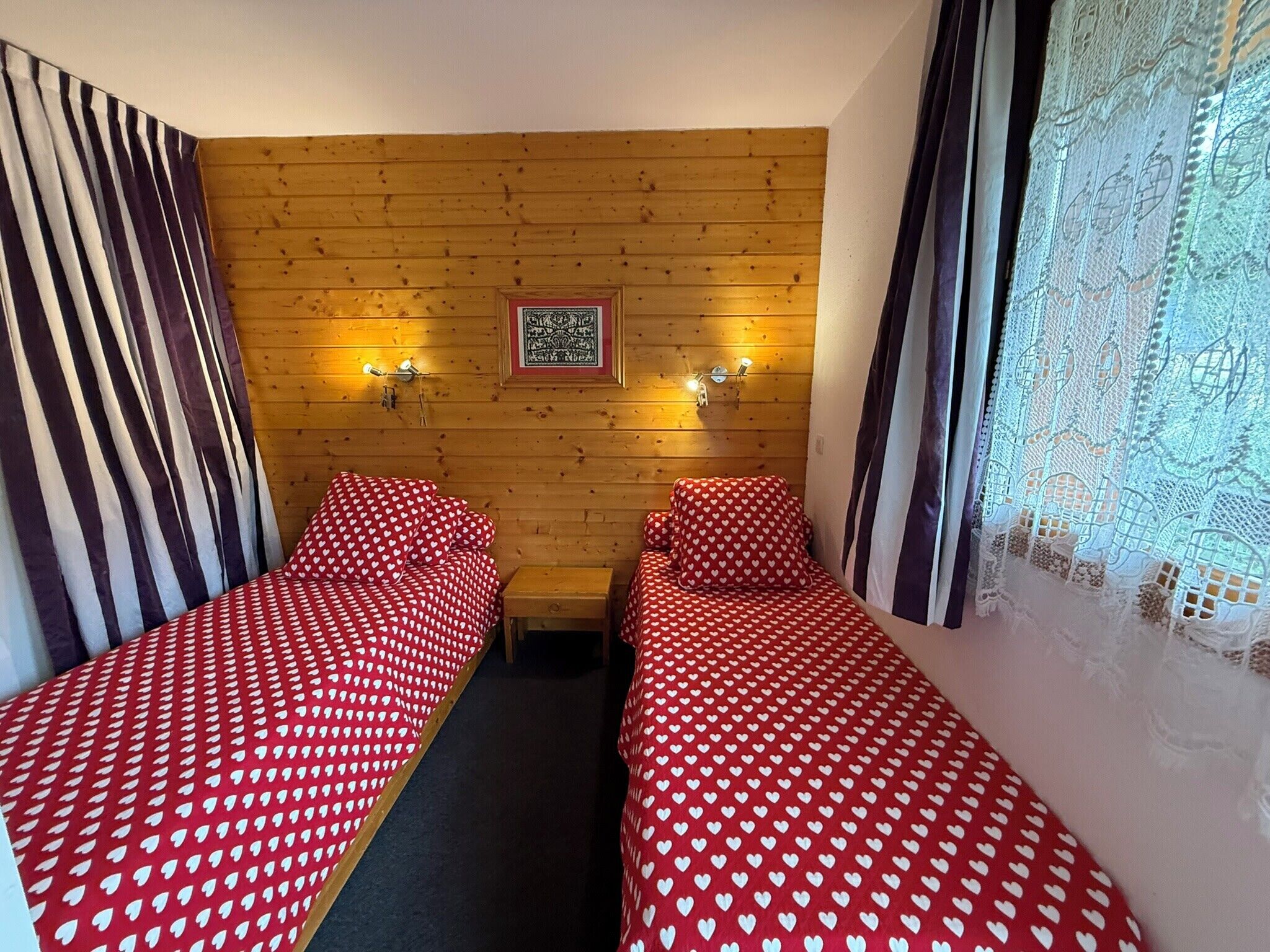 Une chambre avec deux lits simples, des murs en bois, un tableau encadré et une fenêtre ornée de rideaux en dentelle.