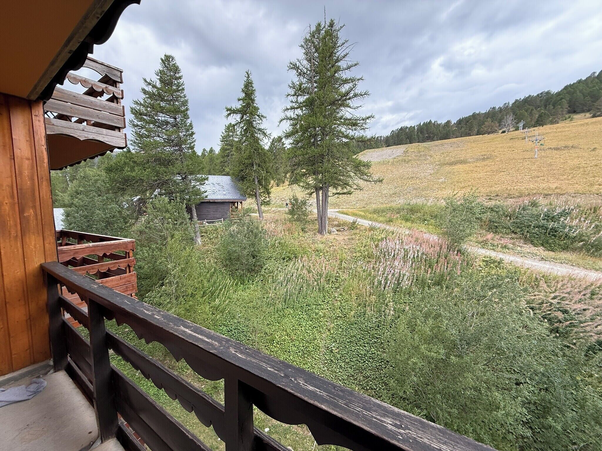 A wooden balcony with a view of a rural landscape, including trees and a small building.
