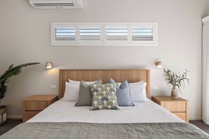 A bedroom with a bed, bedside tables, a wall-mounted air conditioner, a window with blinds, and two wall-mounted lamps. - Coastal Rhythms - A Sophisticated Seaside Retreat (Dunsborough)