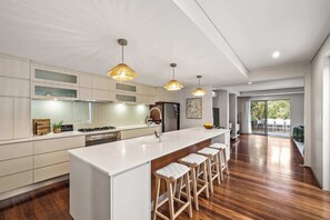 A modern kitchen with a central island, bar stools, pendant lighting, and a view to the outdoors. - Coastal Rhythms - A Sophisticated Seaside Retreat (Dunsborough)