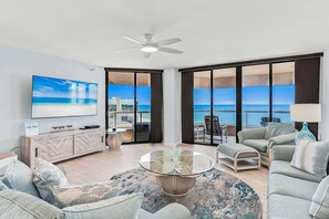 A spacious living room with a large TV, comfortable seating, a glass coffee table, and a view of the ocean. - 🌞CBC 1-15A – Water views from every window! 5 balconies, heated pool, pickleball, gym. (Clearwater Beach)
